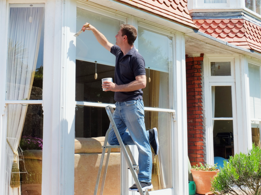 A painter on a ladder applying paint to exterior window trim on a residential home — professional exterior painting services in Manatee County by Razo Painting.